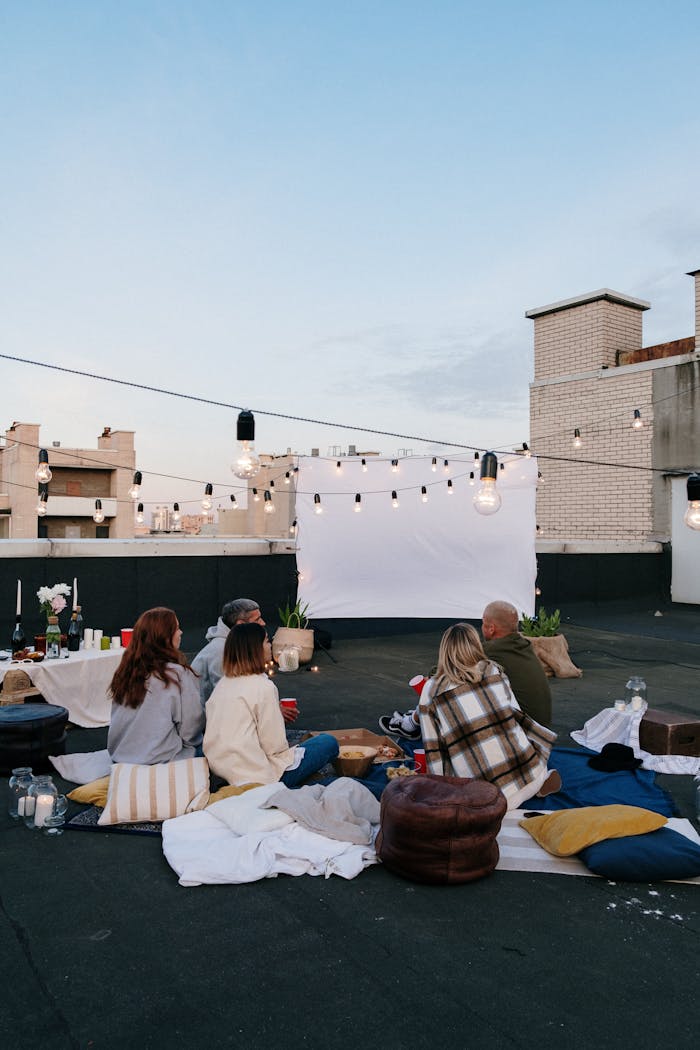 A group of friends enjoys an evening rooftop movie under string lights, sitting on blankets.
