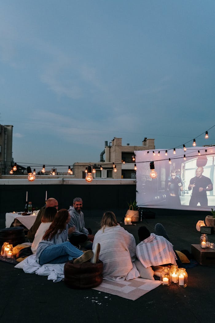 A group of friends enjoying an outdoor movie on a rooftop at dusk, surrounded by lights.