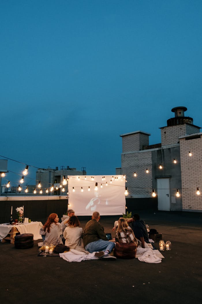 A group of friends enjoys a rooftop movie night under string lights with a projector and cozy blankets.