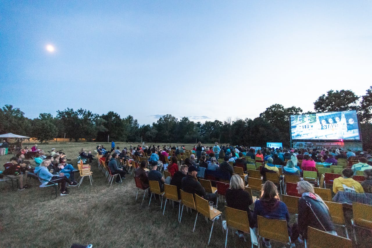 A large group enjoys a movie outdoors at night in a field with a big screen under the moon.